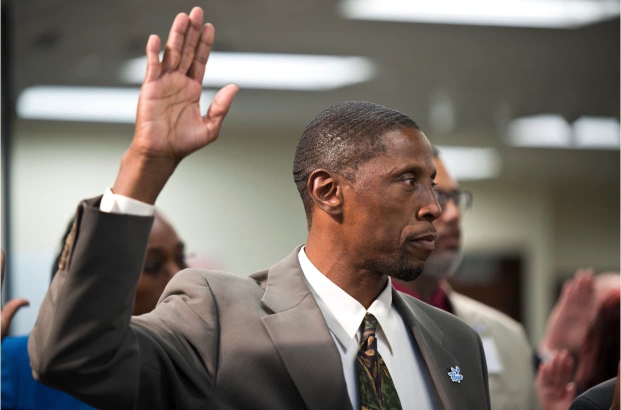 A Black man in a suit raises his right hand while taking an oath.