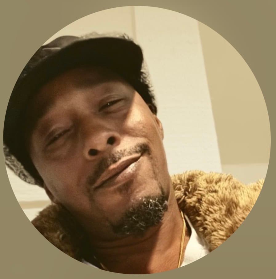 Smiling Black man with a goatee wearing a black cap and a shearling-lined jacket.